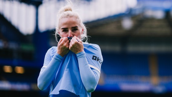 A soccer player in a light blue jersey with a blurred face poses on a football field.