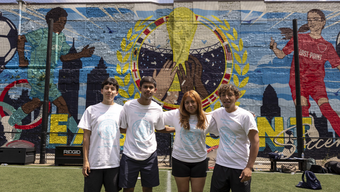 Four young people standing on a soccer field in front of a vibrant mural featuring soccer themes, including a large trophy and players in action.
