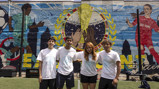 Four young people standing on a soccer field in front of a vibrant mural featuring soccer themes, including a large trophy and players in action.