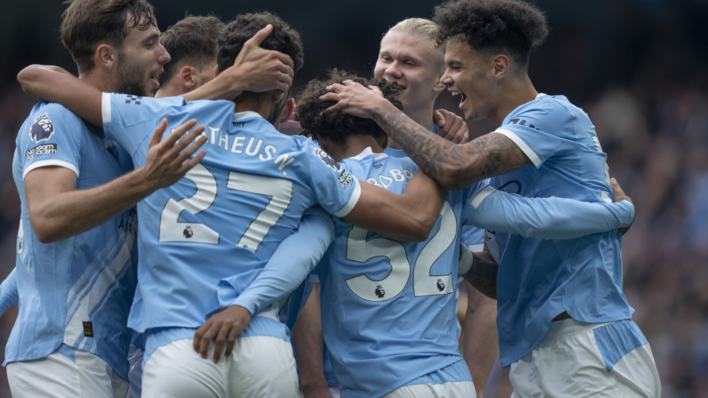 Football players in Manchester City jerseys celebrating on the field, hugging each other after a goal.