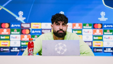A person with a blurred face is speaking at a press conference with a UEFA Champions League backdrop, featuring various sponsor logos.