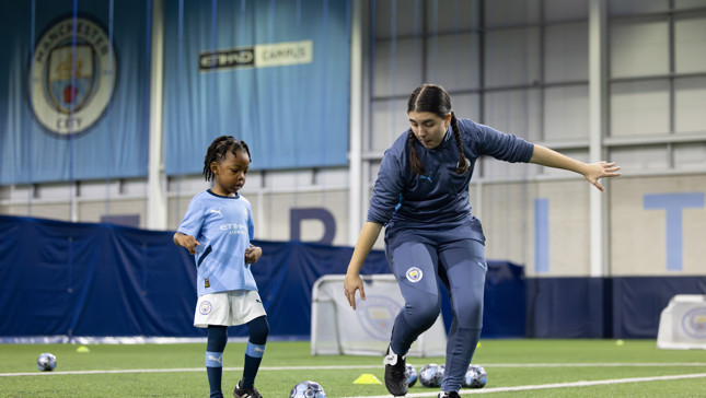 A Manchester City youth player and coach training indoors with footballs. The coach is demonstrating a move while the child observes.