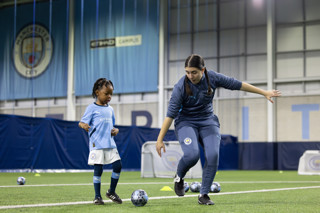 A Manchester City youth player and coach training indoors with footballs. The coach is demonstrating a move while the child observes.