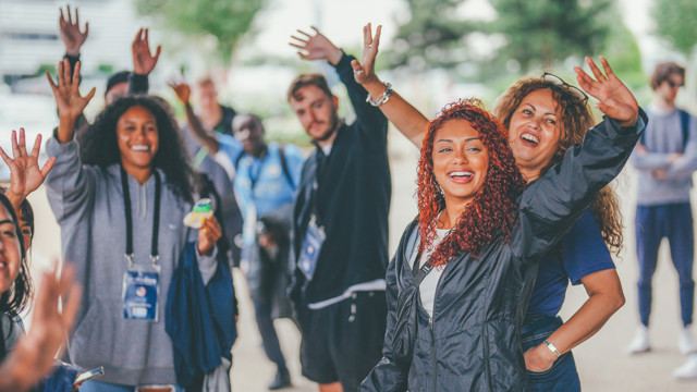 A group of people waving enthusiastically with blurred faces, in an outdoor setting. One person holds a small item, and others have badges hanging from their necks.