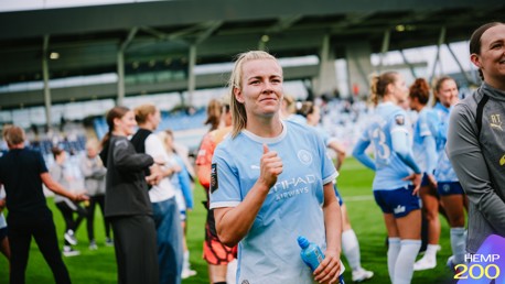 A woman in a Manchester City kit gives a thumbs up gesture on a football field, surrounded by teammates wearing similar kits.