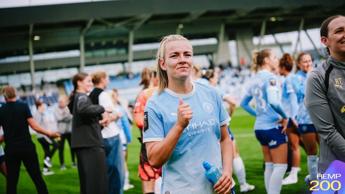 A woman in a Manchester City kit gives a thumbs up gesture on a football field, surrounded by teammates wearing similar kits.