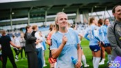A woman in a Manchester City kit gives a thumbs up gesture on a football field, surrounded by teammates wearing similar kits.