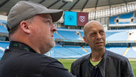 Two people having a conversation inside the Etihad Stadium with a scoreboard showing logos of Manchester City and West Ham in the background.