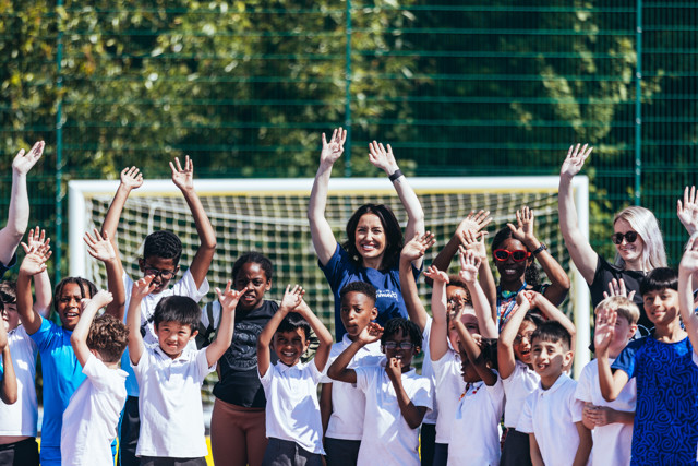 A group of children and adults are raising their hands while standing in front of a soccer goal on a playground. Faces are blurred for privacy.