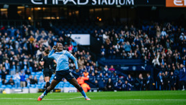 A football player in a Manchester City uniform celebrates a goal on the field at Etihad Stadium with a cheering crowd in the background.