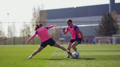 Two soccer players in pink bibs practicing on a field, one attempting a tackle.