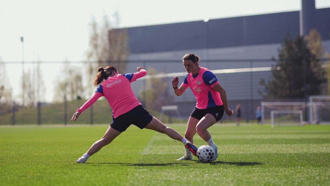 Two soccer players in pink bibs practicing on a field, one attempting a tackle.