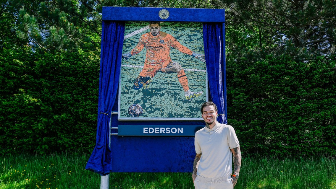 A mosaic display of a Manchester City player named Ederson in a goalkeeper uniform, next to an unidentified person standing, surrounded by green foliage.