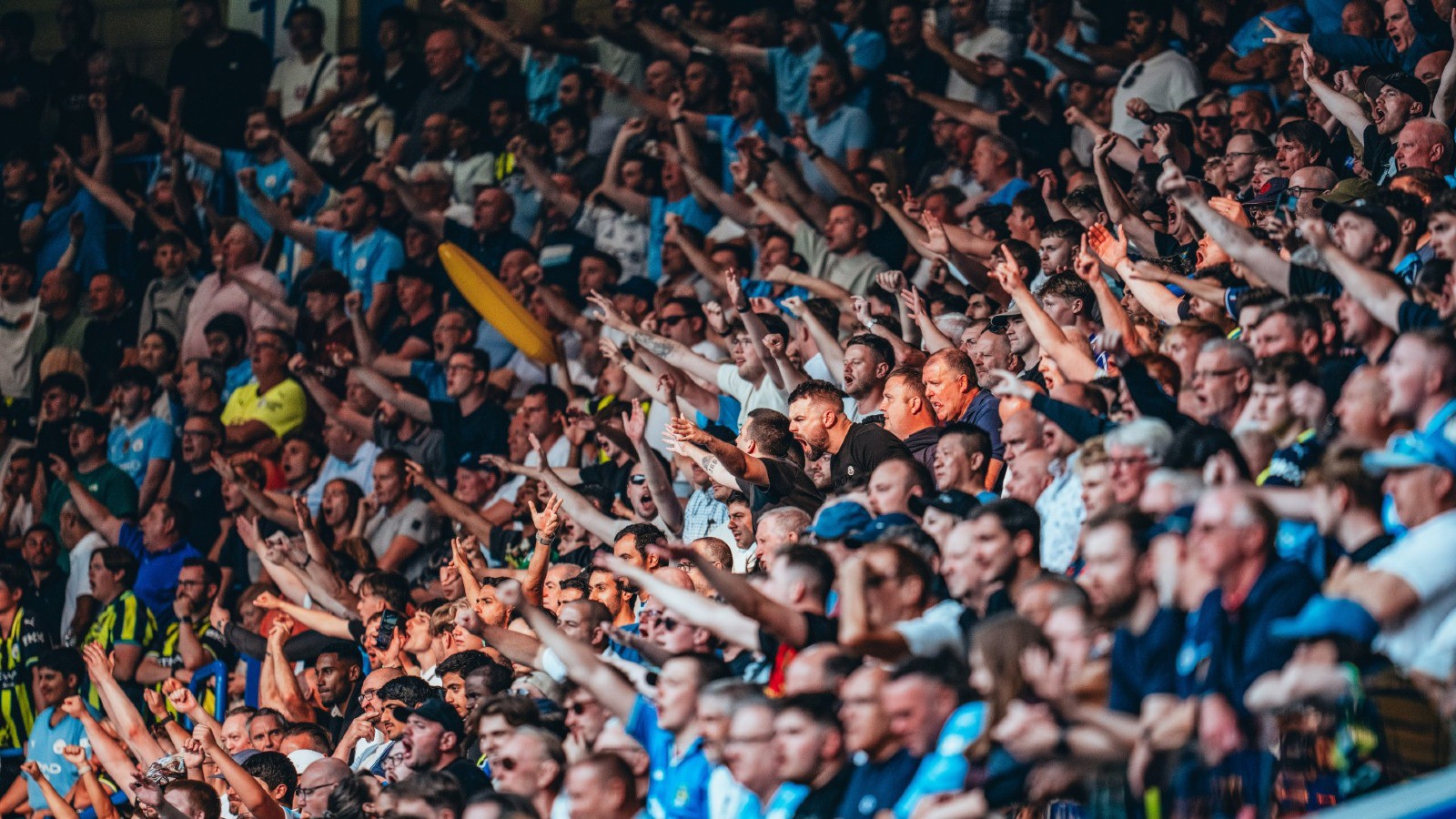 A large crowd of sports fans in a stadium, many wearing blue shirts, enthusiastically cheering and raising their arms.