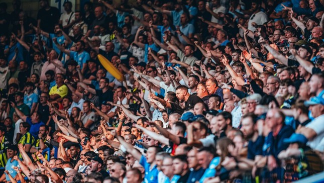 A large crowd of sports fans in a stadium, many wearing blue shirts, enthusiastically cheering and raising their arms.