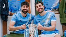 Two Manchester City players in celebratory locker room holding the Carabao Cup trophy with blurred faces.