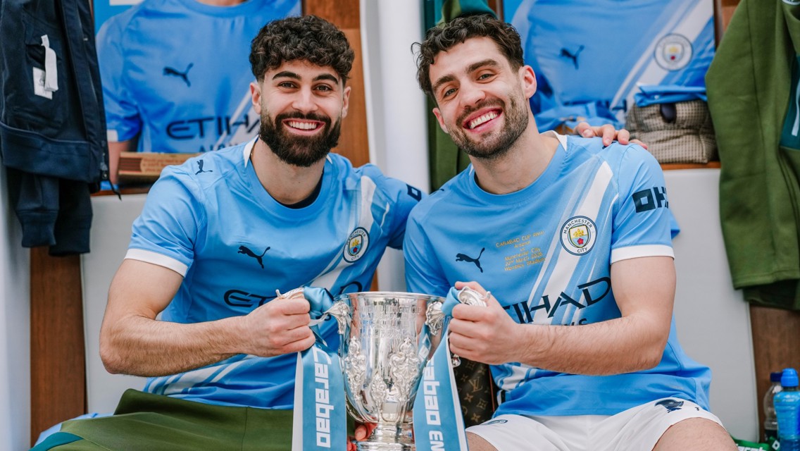 Two Manchester City players in celebratory locker room holding the Carabao Cup trophy with blurred faces.