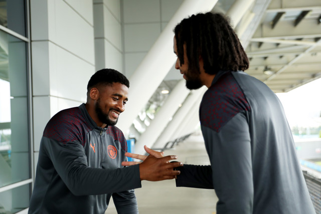 Two Manchester City players are seen shaking hands while wearing training gear with one player's back to the camera and another facing forward with a blurred face.