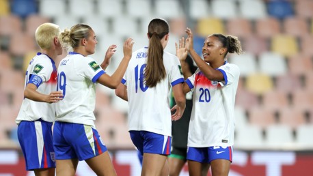 Four women in England football kits celebrating on the field after a match or goal.