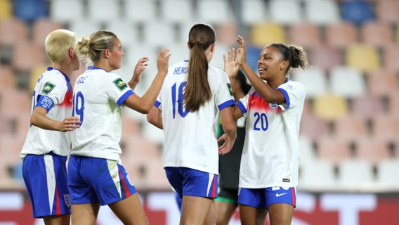 Four women in England football kits celebrating on the field after a match or goal.