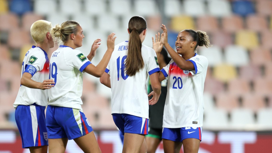 Four women in England football kits celebrating on the field after a match or goal.