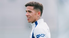 A football player with short hair and tattoos wearing a white jersey with blue accents, looking to the side in a training session.