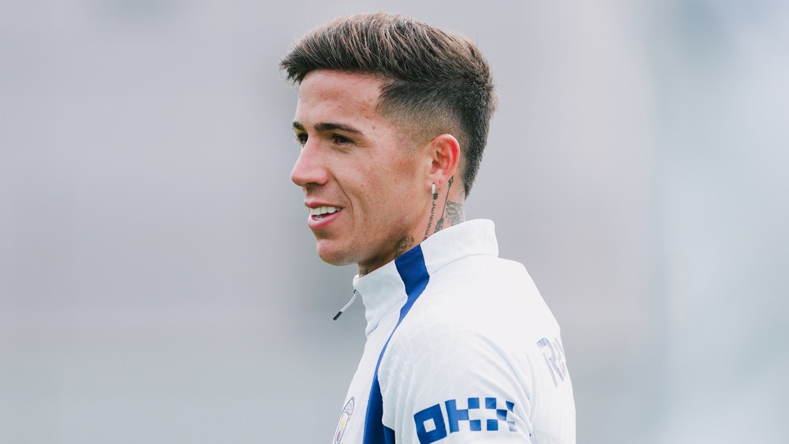 A football player with short hair and tattoos wearing a white jersey with blue accents, looking to the side in a training session.
