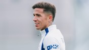 A football player with short hair and tattoos wearing a white jersey with blue accents, looking to the side in a training session.