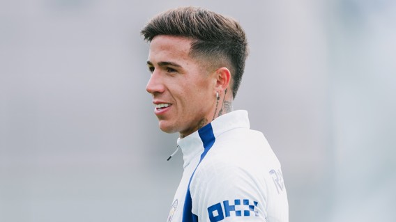 A football player with short hair and tattoos wearing a white jersey with blue accents, looking to the side in a training session.