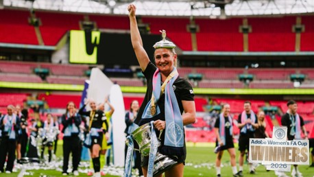 A Manchester City women's player raises the Adobe Women's FA Cup trophy with a team celebrating in the background. The stadium is filled with confetti and fans. The text 'Winners 2026' is prominently displayed.
