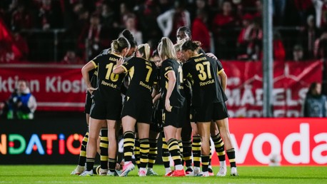 A women's football team in black and yellow jerseys huddles together on the field during a match.