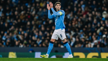 A football player in a light blue Manchester City jersey with the number 5 clapping on the field, surrounded by a crowd.