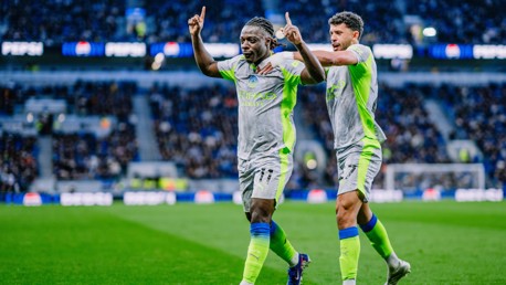 Two soccer players in Manchester City kits celebrate a goal on the field with arms raised.