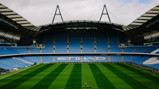 A panoramic view of an empty stadium with blue seats and a well-manicured green field. 'Etihad' is visible on a seating section. The sky appears cloudy.