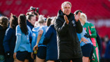 A coach wearing a black jacket claps while a group of female football players in light blue jerseys huddle in the background on a football field.