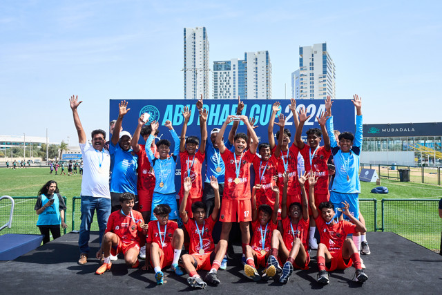 A team in red uniforms celebrates their victory with hands raised at the Abu Dhabi Cup 2023. They pose for a group photo on a podium with medals around their necks.