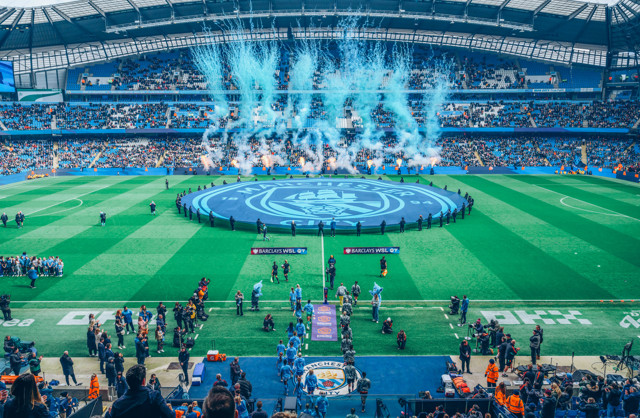 The image shows Manchester City's stadium during a matchday, featuring a large Man City logo banner on the field and blue fireworks in the background as teams enter the pitch with spectators in the stands.