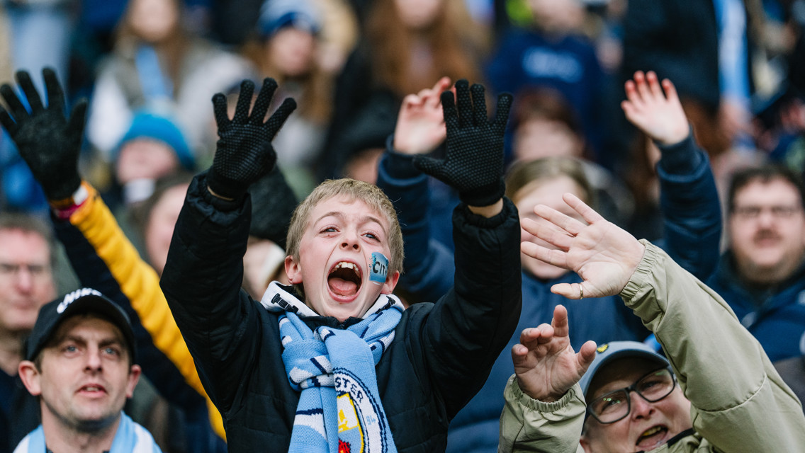 A crowd of fans at a sporting event, with Manchester City scarves visible. The fans are raising their hands and wearing winter clothing.