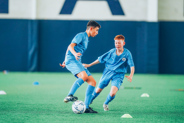 Two young players wearing Manchester City blue kits engage in a training session indoors, with one attempting to dribble past the other.