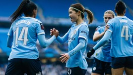 Manchester City women's football players celebrating during a match at the stadium. Players wear blue jerseys, and two of them are shaking hands.