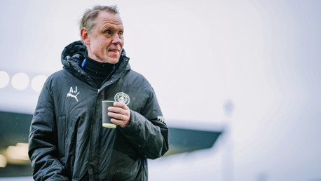 An individual wearing a Manchester City jacket and holding a coffee cup. The jacket features the Puma logo and Manchester City crest.