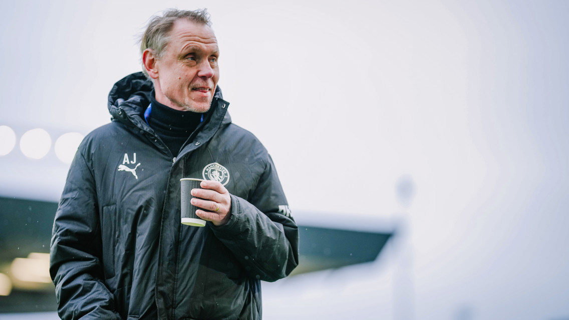 An individual wearing a Manchester City jacket and holding a coffee cup. The jacket features the Puma logo and Manchester City crest.