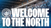 A Manchester City banner with the text 'WELCOME TO THE NORTH' in bold letters against a stadium crowd background showing the Manchester City logo.