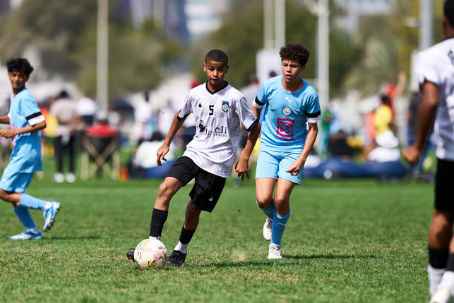 Two young soccer players competing in a match, one in a light blue uniform and the other in a white and black uniform, running on the field with focus on the player in white controlling the ball.