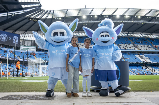 Two children in Manchester City jerseys pose with the team's mascots on a football field at Etihad Stadium.