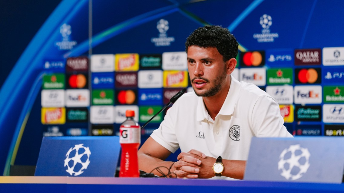 A person in a Manchester City polo speaking at a UEFA Champions League press conference with sponsor logos in the background.