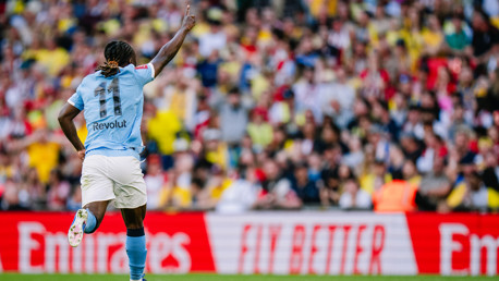 A football player wearing a light blue jersey with '11' and 'Revolut' on the back celebrates a goal in front of a blurred crowd. The scene includes an 'Emirates' advertisement banner.