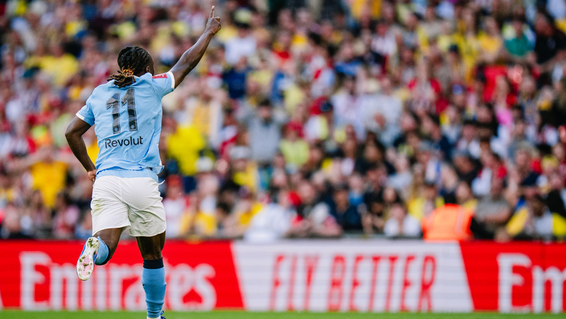 A football player wearing a light blue jersey with '11' and 'Revolut' on the back celebrates a goal in front of a blurred crowd. The scene includes an 'Emirates' advertisement banner.