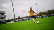 A soccer player in a yellow bib is captured mid-kick on a grassy field during a daytime training session.