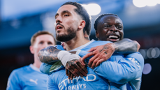 Two football players celebrating wearing Manchester City jerseys, with one player hugging the other.